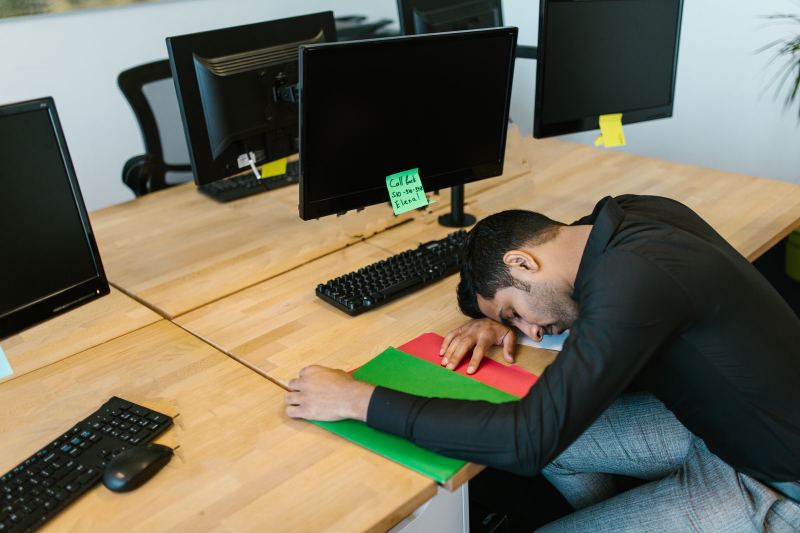 A man in a black shirt rests his head on his arm on a wooden desk, eyes closed, below a monitor with a green sticky note.