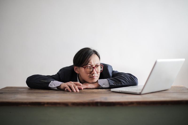 A man in glasses and a dark suit rests his chin on his folded arms at a wooden table, looking at an open laptop.
