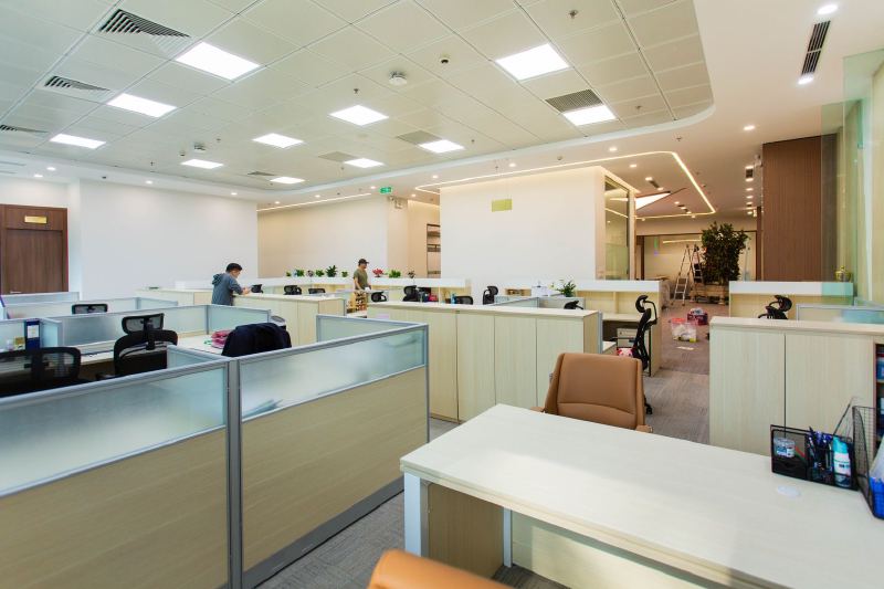 A wide open-plan office with rows of low cubicles under ceiling lights, and two people far off at the back.