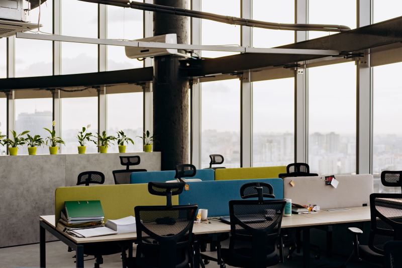 An empty open-plan office with bench desks, mesh chairs, green, blue and gray desk dividers, potted plants and floor-to-ceiling windows over a city.