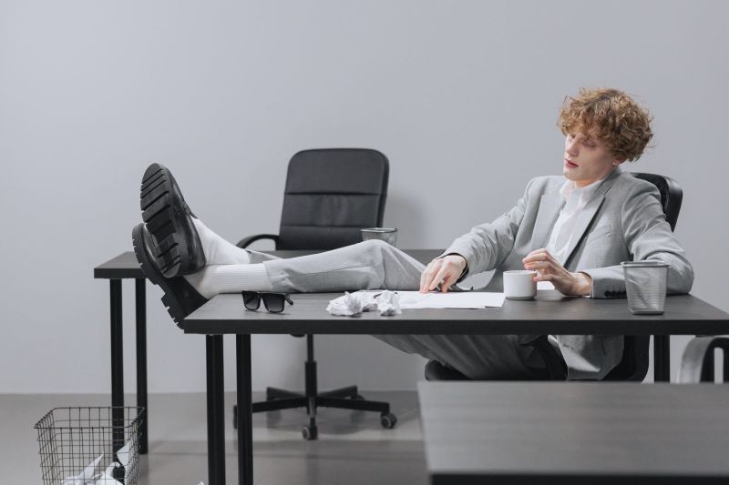 A young man in a gray suit leans back with his shoes on the desk, holding a cup, beside crumpled paper and sunglasses.