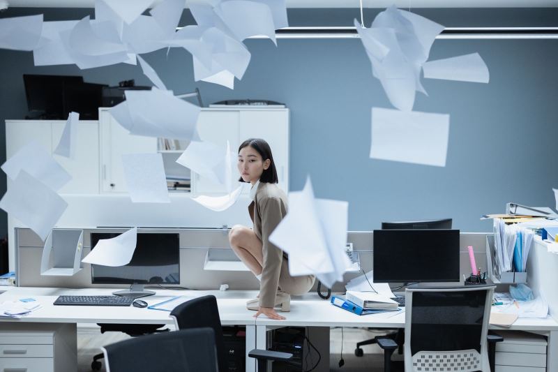 A woman crouches on top of an office desk while sheets of paper fly through the air around her.