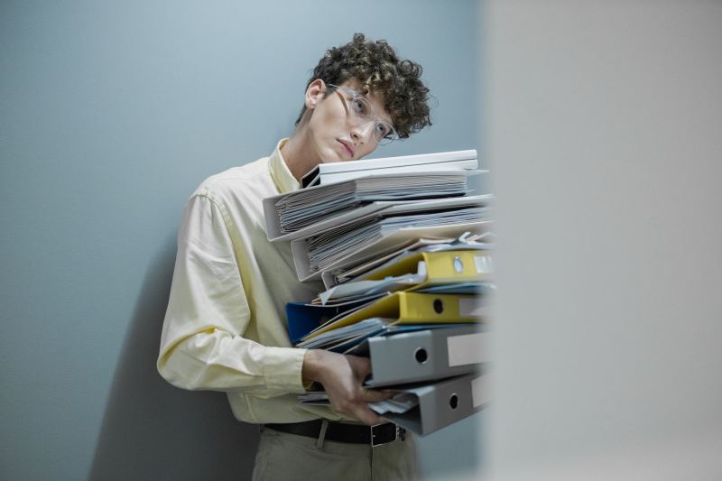 A young man in glasses leans against a wall, carrying a tall stack of ring binders and folders.