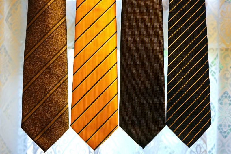 Four neckties in brown, orange, dark brown and black with gold stripes hang in front of a lace curtain.