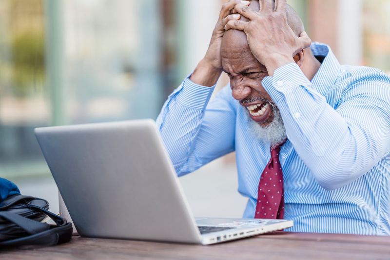 An older man in a blue striped shirt and red tie grips his head with both hands and grimaces at a laptop.