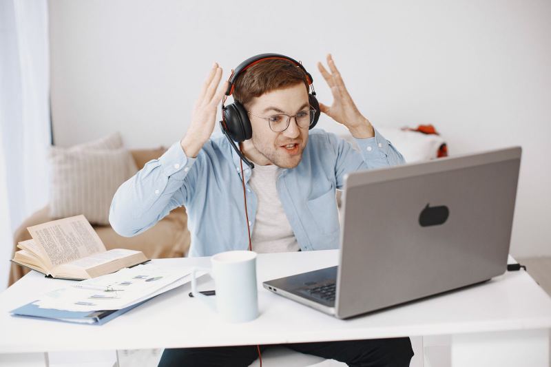 A man in headphones and glasses raises both hands at a laptop on a white desk, next to a mug and printed charts.