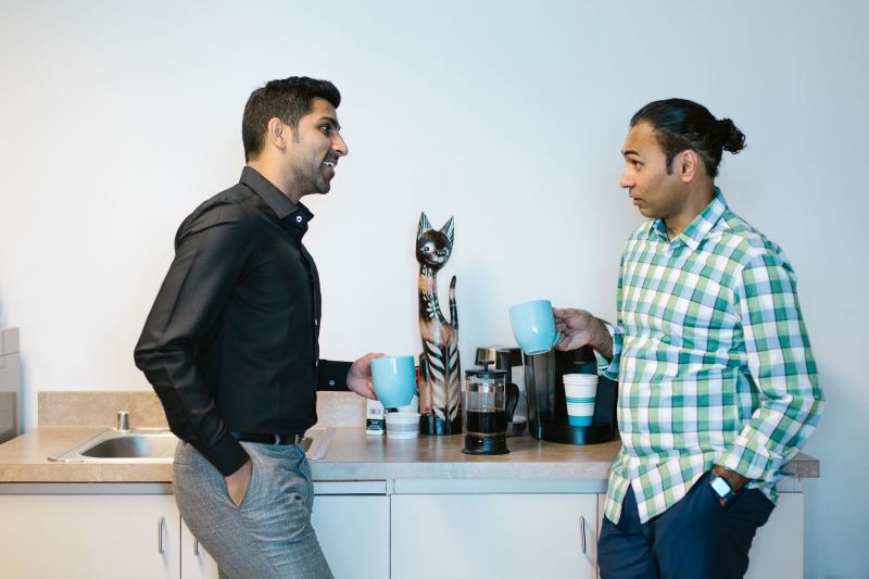 Two men holding blue mugs talk beside a coffee maker, a French press and a wooden cat figure in an office kitchenette.