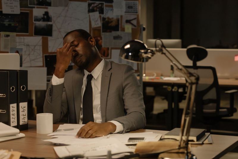 A man in a gray suit and tie pinches the bridge of his nose at a desk covered in papers, with a desk lamp, a mug and numbered binders, in a dark office with a corkboard of photos and red string behind him.