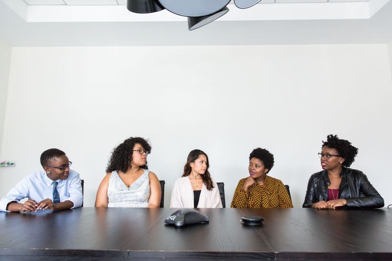 Five people sit along one side of a long dark conference table, with a conference phone in front of them and a bare white wall behind.