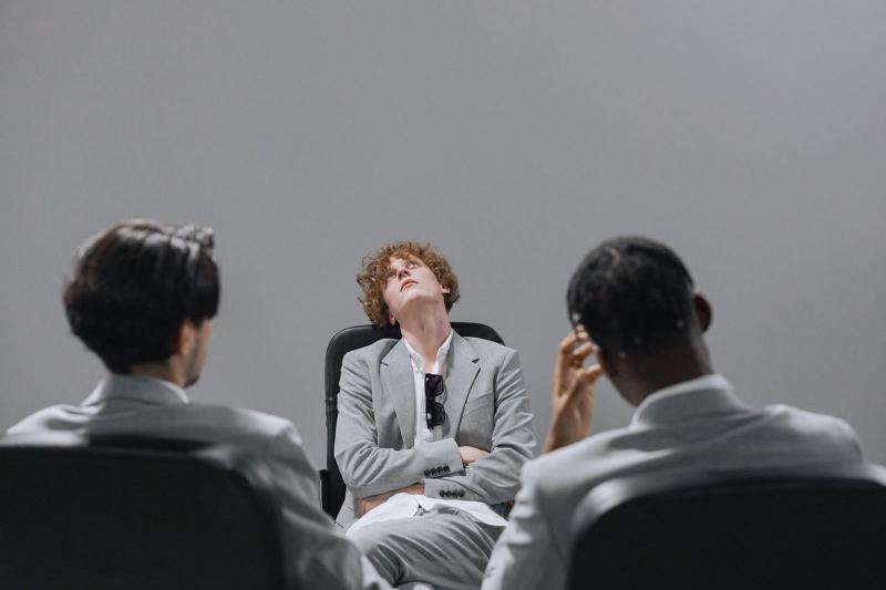 A man in a gray suit sits back in his chair with his arms crossed, looking up, while two colleagues sit with their backs to the camera.