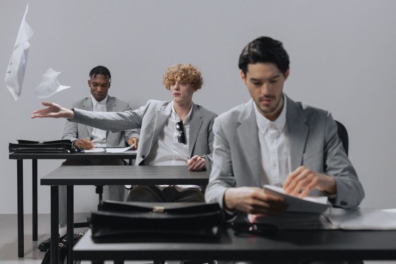 Three men in gray suits sit at desks, and one tosses sheets of paper into the air while the other two keep working.