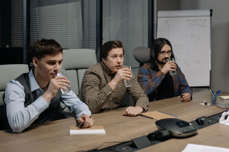 Three men at a conference table with glasses of water, two of them drinking while the one in the middle talks, with a conference phone in front and a flipchart graph behind them.