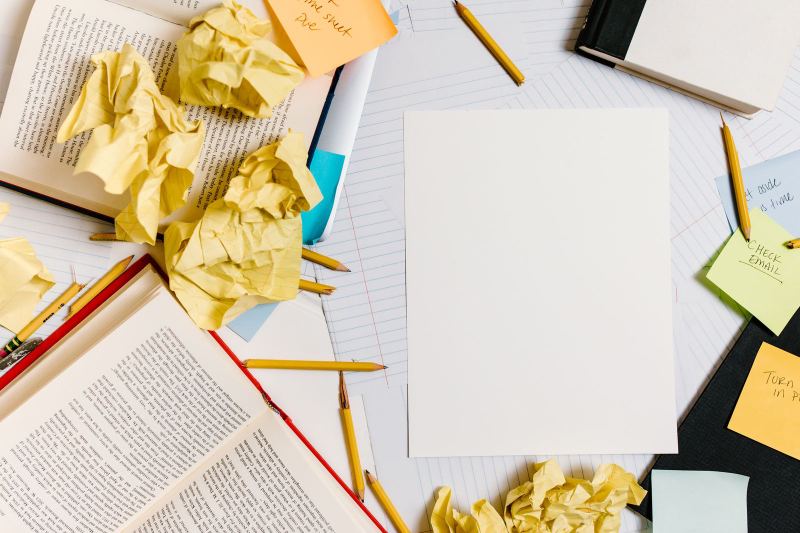 Overhead view of a desk covered in crumpled yellow paper, open books, pencils and sticky notes, one saying Check email, with a blank sheet in the middle.