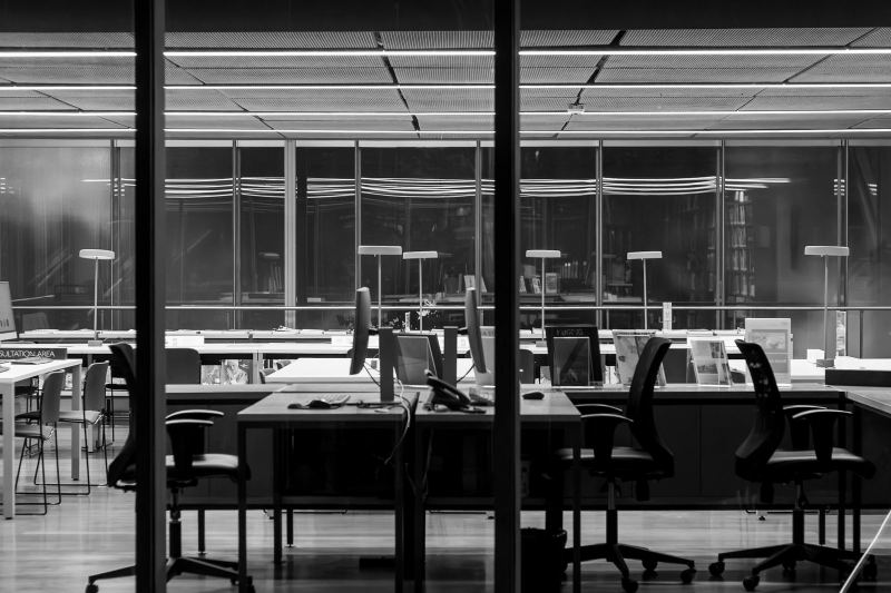 Black-and-white view through a glass wall into an empty office at night, with rows of desks, office chairs, monitors and desk lamps.