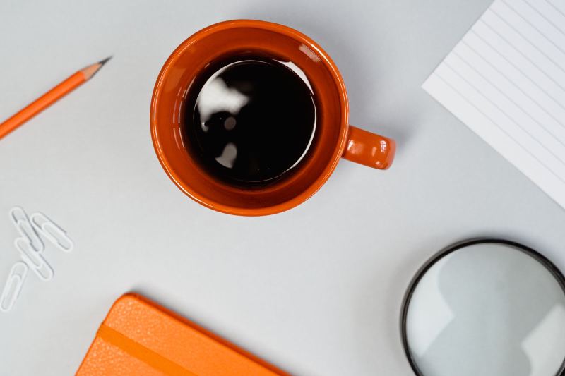 Overhead view of an orange mug of black coffee on a gray desk, with an orange pencil, paper clips, an orange notebook, a lined card and a magnifying glass.