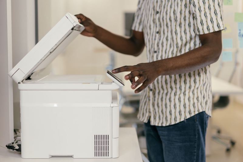 A person in a striped short-sleeved shirt lifts the lid of a white photocopier and taps its small touch screen.