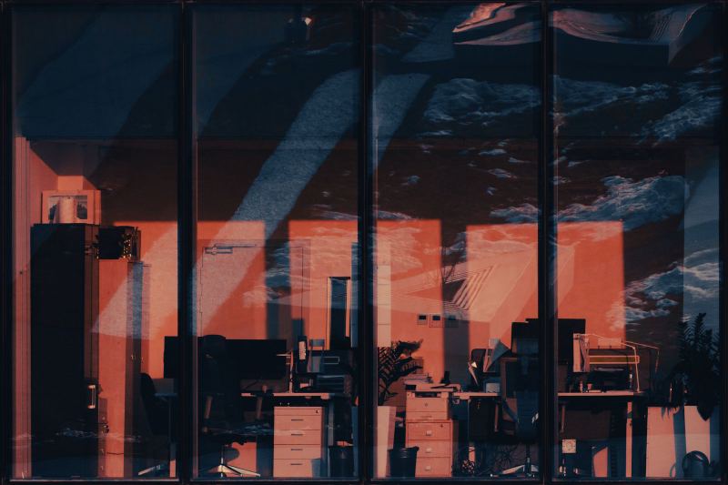 Empty office desks, drawers and chairs seen through large windows at dusk, the room lit red and orange, the sky reflected in the glass.