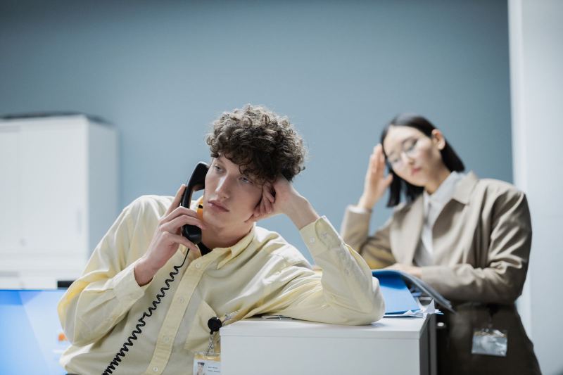 A young man in a yellow shirt leans on a filing cabinet with a corded desk phone to his ear, while a woman behind him rests her head on her hand.