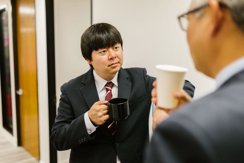 A man in a dark suit and red striped tie holds a black mug while a colleague, seen from behind, holds a paper cup.