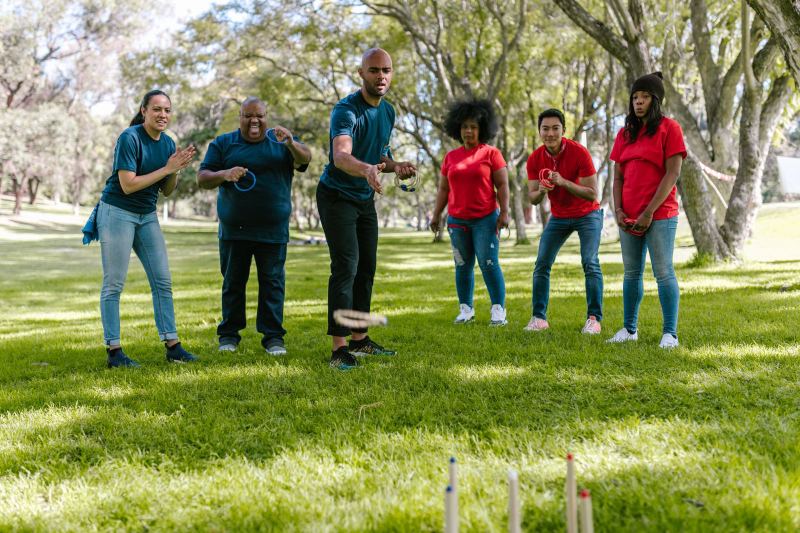 Six people in blue and red T-shirts play ring toss on a lawn under trees.