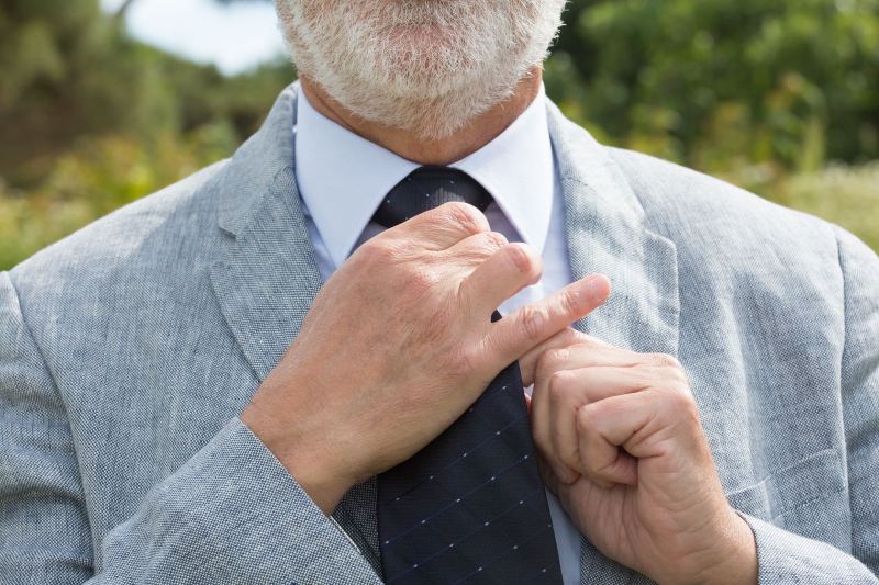 Close-up of a gray-bearded man in a light gray jacket straightening a navy tie outdoors, cropped at the mouth.