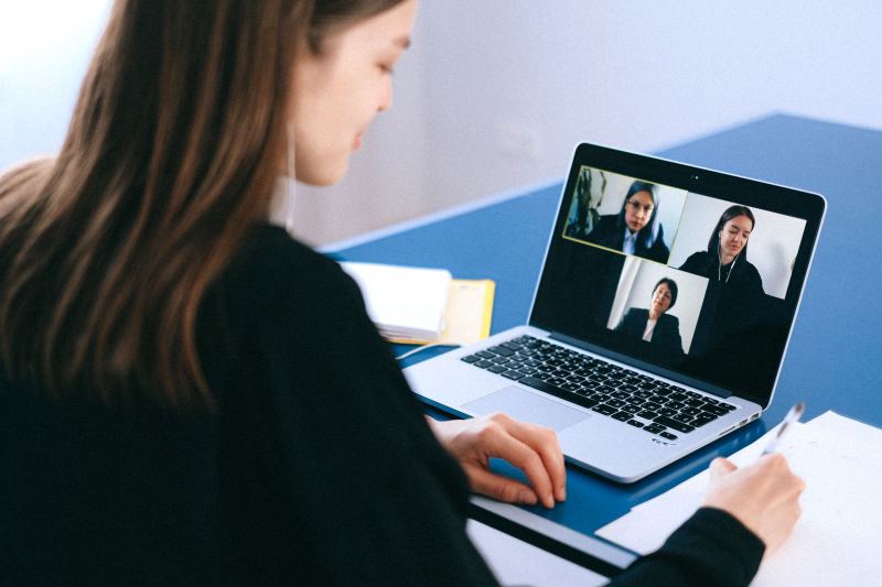 A woman seen from behind takes notes beside a laptop showing a video call with three people in a grid.