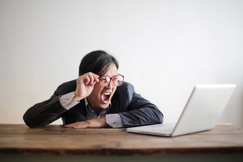 A man in a suit and red glasses leans on a wooden table with his mouth wide open, lifting his glasses at a laptop.